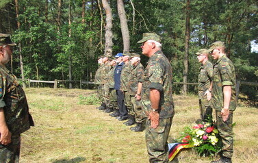 Grupa os&oacute;b w wojskowych, maskujących mundurach stoi w szeregu na leśnej polanie podczas uroczystości. Przed nimi znajduje się wieniec z kwiat&oacute;w z wstęgą w kolorach flagi Niemiec. Wszyscy stoją na baczność, zwr&oacute;ceni w stronę pojedynczej osoby w mundurze, k