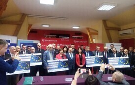 Grupa os&oacute;b pozuje do zdjęcia w sali konferencyjnej podczas oficjalnego wydarzenia. Kilka z nich trzyma duże symboliczne czeki z motywem flagi Unii Europejskiej i kwotami dofinansowania. W tle stoją czerwone plansze z napisem &bdquo;25. Mazowsze&rdquo;. W kadrze widoc