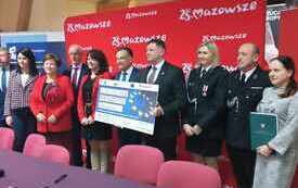 Dziesięć os&oacute;b stoi na tle czerwonej planszy z napisem &bdquo;25 lat Mazowsze&rdquo;. Wszyscy są ubrani formalnie, część w garniturach, a jedna osoba w mundurze. Osoba w centrum trzyma duży symboliczny czek z flagą Unii Europejskiej i kwotą &bdquo;1 253 782,00 zł&rdquo;, dotycząc