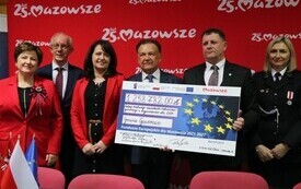 Sześć os&oacute;b stoi na tle czerwonej planszy z napisem &bdquo;25 lat Mazowsze&rdquo;. Dwie osoby trzymają duży symboliczny czek z flagą Unii Europejskiej i kwotą &bdquo;1 258 782,00 zł&rdquo;, dotyczącym zakupu średniego samochodu ratowniczo‑gaśniczego dla OSP w gminie Goworowo. Wsz
