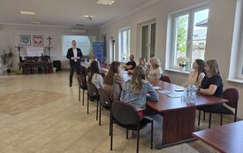 Zdjęcie przedstawia spotkanie w jasnej sali z dużymi oknami. Przy prostokątnym drewnianym stole siedzi grupa os&oacute;b słuchających mężczyzny stojącego z przodu, ubranego w formalny str&oacute;j. Za nim znajduje się ekran z prezentacją oraz dwa herby i krzyż na ścian