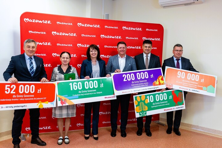 Zdjęcie przedstawia sześć os&oacute;b stojących w&nbsp;pomieszczeniu na&nbsp;tle czerwonej ścianki z&nbsp;powtarzającym się białym napisem &bdquo;Mazowsze&rdquo;. Każda z&nbsp;os&oacute;b trzyma duży, kolorowy czek ceremonialny z&nbsp;nadrukowaną kwotą oraz&nbsp;opisem przeznaczenia środk&oacute;w dla Gminy Goworowo. Kwoty na&nbsp;czekach są r&oacute;żne i&nbsp;dotyczą m.in. modernizacji strażnicy OSP, przebudowy dr&oacute;g, zakupu stroj&oacute;w ludowych oraz&nbsp;zakupu samochodu dla jednostki OSP. Osoby ustawione są w&nbsp;jednym rzędzie, zwr&oacute;cone w&nbsp;stronę aparatu, pozują do zdjęcia i&nbsp;prezentują otrzymane czeki. Całość ma charakter oficjalnego wydarzenia związanego z&nbsp;przekazaniem dofinansowań na&nbsp;lokalne projekty.