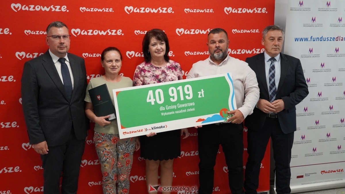 Zdjęcie przedstawia pięć os&oacute;b stojących w&nbsp;rzędzie na&nbsp;tle czerwonej ścianki z&nbsp;powtarzającym się białym napisem &bdquo;Mazowsze&rdquo;. Wszyscy pozują do fotografii, trzymając wsp&oacute;lnie duży, prostokątny czek ceremonialny. Na czeku widnieje kwota &bdquo;40 919 zł&rdquo;, informacja o&nbsp;przyznaniu środk&oacute;w dla Gminy Goworowo oraz&nbsp;opis celu: &bdquo;Wykonanie nasadzeń zieleni&rdquo;. W dolnej części czeku znajduje się napis &bdquo;Mazowsze dla klimatu&rdquo;. Osoby na&nbsp;zdjęciu są ubrane odświętnie, a&nbsp;całość ma charakter oficjalnego wydarzenia związanego z&nbsp;przekazaniem dofinansowania na&nbsp;działania proekologiczne.