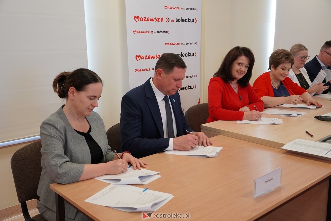 Zdjęcie przedstawia sześć os&oacute;b siedzących przy prostokątnym, drewnianym stole w&nbsp;sali o&nbsp;neutralnych, jasnych ścianach. Każda z&nbsp;os&oacute;b ma przed sobą dokumenty, a&nbsp;część z&nbsp;nich podpisuje lub&nbsp;przegląda papiery. W tle widoczna jest czerwona ścianka z&nbsp;powtarzającym się białym napisem &bdquo;Mazowsze dla sołectw&rdquo;. Uczestnicy są ubrani formalnie, co podkreśla oficjalny charakter spotkania. Na stole leżą długopisy oraz&nbsp;tabliczka z&nbsp;nazwą instytucji lub&nbsp;uczestnika. Scena przedstawia moment podpisywania dokument&oacute;w w&nbsp;ramach lokalnego projektu lub&nbsp;inicjatywy samorządowej.