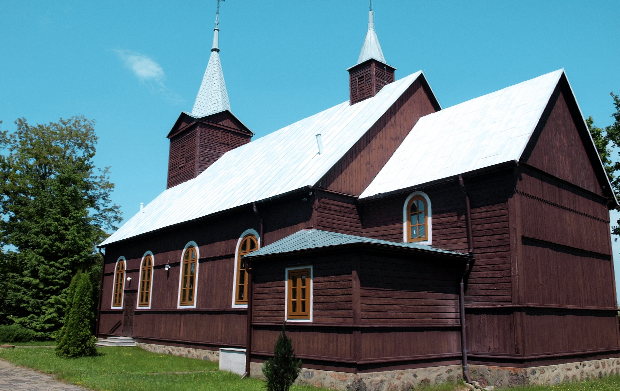 Zdjęcie przedstawia drewniany kości&oacute;ł o&nbsp;tradycyjnej, historycznej architekturze, stojący na&nbsp;otwartej, zielonej przestrzeni. Budowla ma ciemne, brązowe ściany wykonane z&nbsp;pionowych desek oraz&nbsp;wysokie, strome dachy pokryte jasną, metaliczną blachą. Z gł&oacute;wnego dachu wyrastają dwie smukłe wieże zakończone ostro zakończonymi hełmami, na&nbsp;kt&oacute;rych umieszczono metalowe krzyże.  Fasada kościoła jest ozdobiona szeregiem wysokich, wąskich okien o&nbsp;łukowatych zwieńczeniach. Ramy okien są białe, co kontrastuje z&nbsp;ciemnym drewnem ścian. Z prawej strony widoczna jest mniejsza przybud&oacute;wka z&nbsp;osobnym, niższym dachem, r&oacute;wnież pokrytym metalową blachą. Cała konstrukcja stoi na&nbsp;solidnej, kamiennej podmur&oacute;wce.  Kości&oacute;ł otacza zadbana, zielona trawa, a&nbsp;w tle rosną drzewa o&nbsp;gęstych koronach. Niebo jest czyste i&nbsp;intensywnie błękitne, bez chmur, co sugeruje pogodny, słoneczny dzień. Całość tworzy spokojną, wiejską scenerię, podkreślając tradycyjne rzemiosło i&nbsp;charakterystyczny styl drewnianych świątyń.