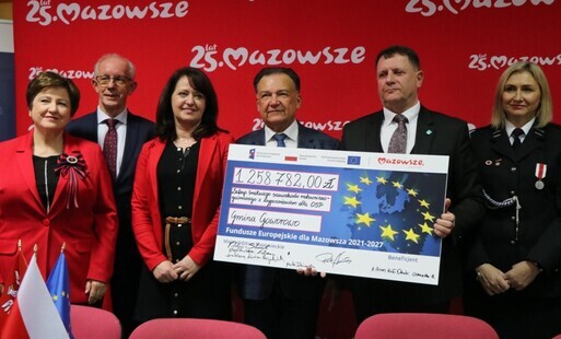 Sześć osób stoi na tle czerwonej planszy z napisem „25 lat Mazowsze”. Dwie osoby trzymają duży symboliczny czek z flagą Unii Europejskiej i kwotą „1 258 782,00 zł”, dotyczącym zakupu średniego samochodu ratowniczo‑gaśniczego dla OSP w gminie Goworowo. Wsz