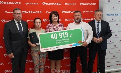 Zdjęcie przedstawia pięć osób stojących w rzędzie na tle czerwonej ścianki z powtarzającym się białym napisem „Mazowsze”. Wszyscy pozują do fotografii, trzymając wspólnie duży, prostokątny czek ceremonialny. Na czeku widnieje kwota „40 919 zł”, informacja