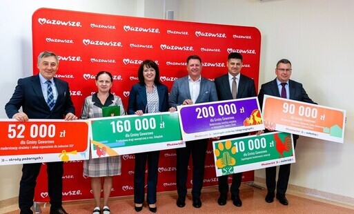 Zdjęcie przedstawia sześć osób stojących w pomieszczeniu na tle czerwonej ścianki z powtarzającym się białym napisem „Mazowsze”. Każda z osób trzyma duży, kolorowy czek ceremonialny z nadrukowaną kwotą oraz opisem przeznaczenia środków dla Gminy Goworowo.