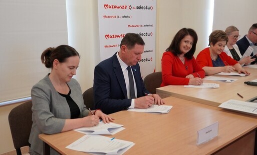 Zdjęcie przedstawia sześć osób siedzących przy prostokątnym, drewnianym stole w sali o neutralnych, jasnych ścianach. Każda z osób ma przed sobą dokumenty, a część z nich podpisuje lub przegląda papiery. W tle widoczna jest czerwona ścianka z powtarzający