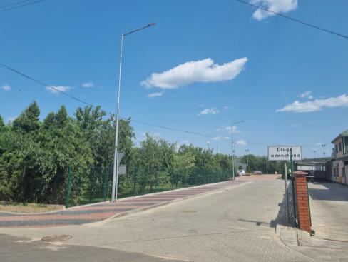 Zdjęcie przedstawia prostą, świeżo wyremontowaną drogę asfaltową biegnącą przez spokojną okolicę. Po lewej stronie znajduje się chodnik oddzielony zielonym ogrodzeniem, za którym rośnie gęsta roślinność. Wzdłuż jezdni ustawione są wysokie latarnie uliczne