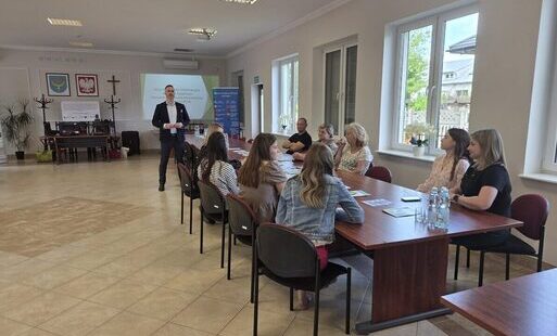 Zdjęcie przedstawia spotkanie w jasnej sali z dużymi oknami. Przy prostokątnym drewnianym stole siedzi grupa osób słuchających mężczyzny stojącego z przodu, ubranego w formalny strój. Za nim znajduje się ekran z prezentacją oraz dwa herby i krzyż na ścian