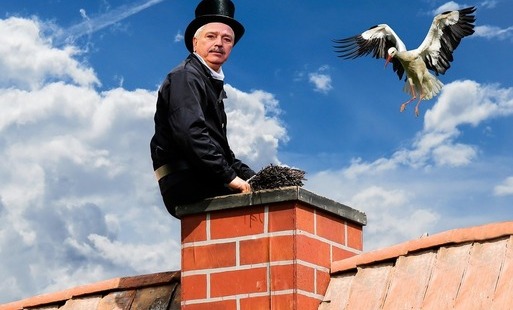 Zdjęcie przedstawia kominiarza w czarnym stroju i wysokim kapeluszu, siedzącego na murowanym kominie na dachu. Obok komina leci bocian z rozpostartymi skrzydłami, jakby miał za chwilę wylądować. W tle widać niebieskie niebo z białymi chmurami.