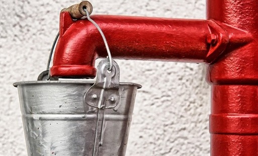 Zdjęcie przedstawia czerwony metalowy kran lub rurę zakończoną wylewką, na której wisi małe, srebrne metalowe wiaderko zawieszone na drucianym uchwycie. W tle znajduje się jasna, chropowata ściana.