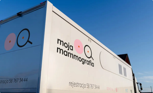 Mobilna pracownia mammograficzna w formie białego samochodu ciężarowego stoi na zewnątrz w słoneczny dzień. Na burcie widoczny jest napis „moja mammografia” oraz grafiki przypominające lupy na okrągłych kształtach, symbolizujące badanie piersi. Pod spodem