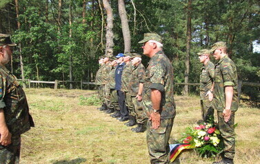 Grupa os&oacute;b w wojskowych, maskujących mundurach stoi w szeregu na leśnej polanie podczas uroczystości. Przed nimi znajduje się wieniec z kwiat&oacute;w z wstęgą w kolorach flagi Niemiec. Wszyscy stoją na baczność, zwr&oacute;ceni w stronę pojedynczej osoby w mundurze, k