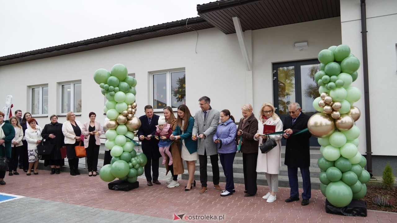 Zdjęcie przedstawia uroczyste przecięcie wstęgi przed nowym budynkiem o&nbsp;jasnej elewacji i&nbsp;ciemnym dachu, co symbolizuje jego oficjalne otwarcie. Na pierwszym planie stoi grupa os&oacute;b w&nbsp;eleganckich strojach, ustawionych w&nbsp;szeregu między dwoma dekoracyjnymi kolumnami z&nbsp;zielonych i&nbsp;złotych balon&oacute;w. Każda z&nbsp;os&oacute;b trzyma fragment zielonej wstęgi, kt&oacute;rą wsp&oacute;lnie przecinają. Wśr&oacute;d uczestnik&oacute;w widać kobiety i&nbsp;mężczyzn w&nbsp;r&oacute;żnym wieku, a&nbsp;jedna z&nbsp;kobiet trzyma małe dziecko. W tle znajduje się wejście do budynku z&nbsp;przeszklonymi drzwiami i&nbsp;kilkoma oknami, a&nbsp;po lewej stronie grupy stoją inni goście obserwujący wydarzenie. Na dole zdjęcia widoczny jest napis &bdquo;eostroleka.pl&rdquo;, wskazujący źr&oacute;dło fotografii. Scena ma charakter oficjalny i&nbsp;uroczysty, prawdopodobnie dokumentuje otwarcie instytucji publicznej lub&nbsp;lokalnego obiektu społecznego.