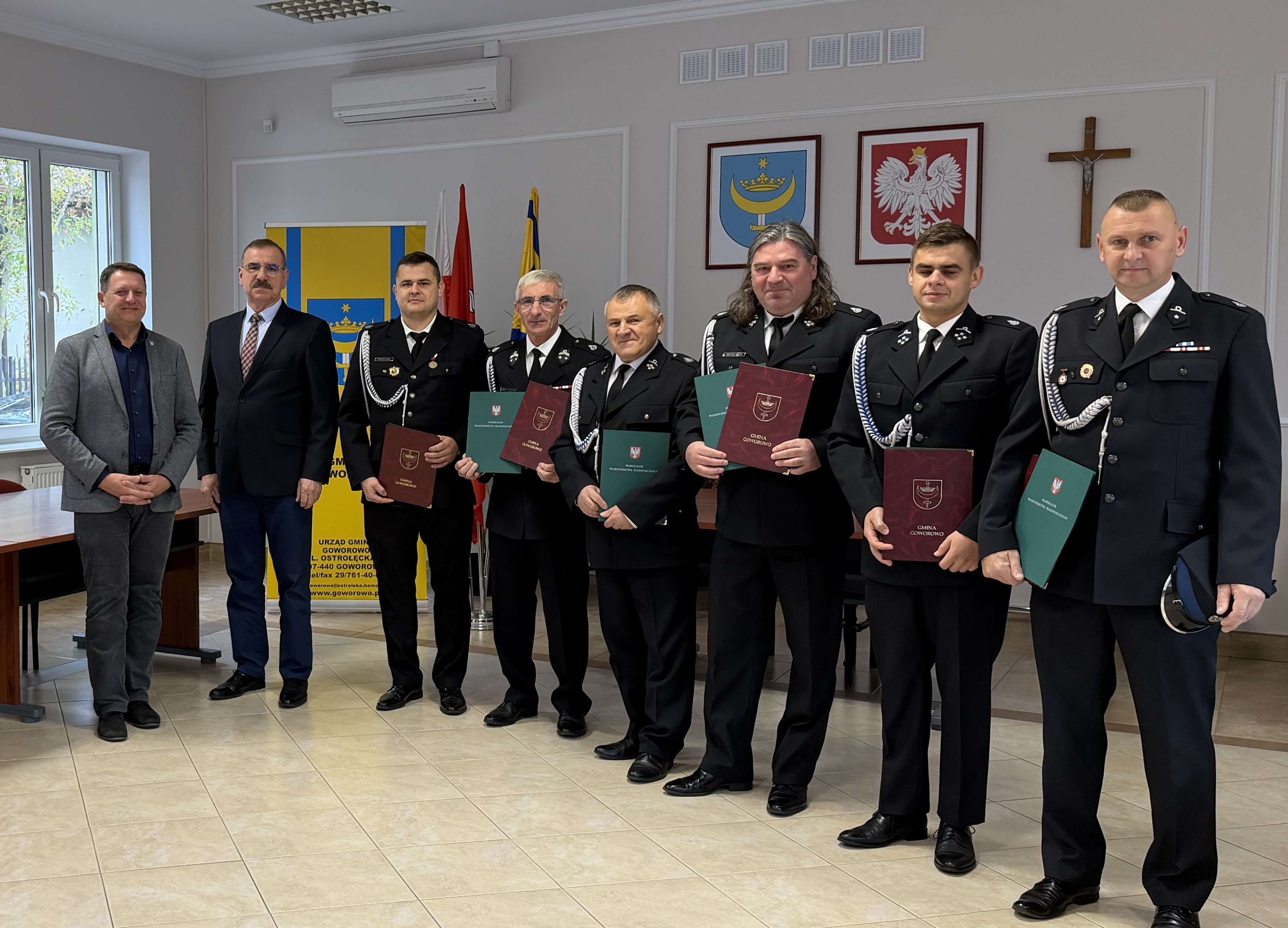 Grupa umundurowanych strażak&oacute;w-ochotnik&oacute;w i&nbsp;dw&oacute;ch cywilnych mężczyzn w&nbsp;sali urzędu trzymających certyfikaty, w&nbsp;tle flagi i&nbsp;godła państwowe.