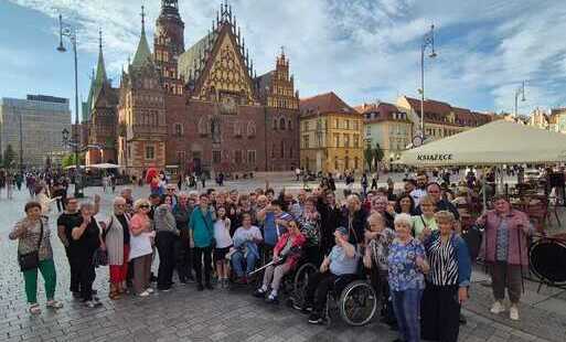 Grupa uczestników na wrocławskim rynku przed zabytkowym ratuszem, niektórzy z wózkami inwalidzkimi, obok namiot handlowy i stoiska pod baldachimem.