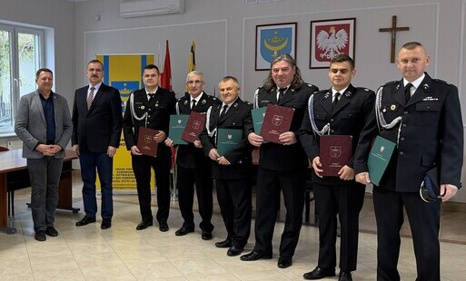 Grupa umundurowanych strażaków-ochotników i dwóch cywilnych mężczyzn w sali urzędu trzymających certyfikaty, w tle flagi i godła państwowe.