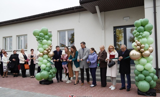 Grupa osób stoi przed budynkiem przedszkola, przecinając pamiątkową wstęgę, a po bokach stoją duże dekoracje z zielonych i złotych balonów.