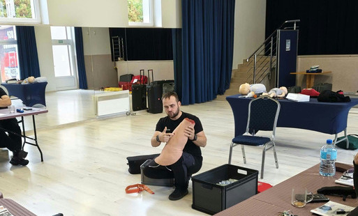 Mężczyzna siedzi na podłodze w sali szkoleniowej i pokazuje dłonie, na pierwszym planie stoją krzesło i czarna skrzynia, w tle sprzęt i niebieskie zasłony.